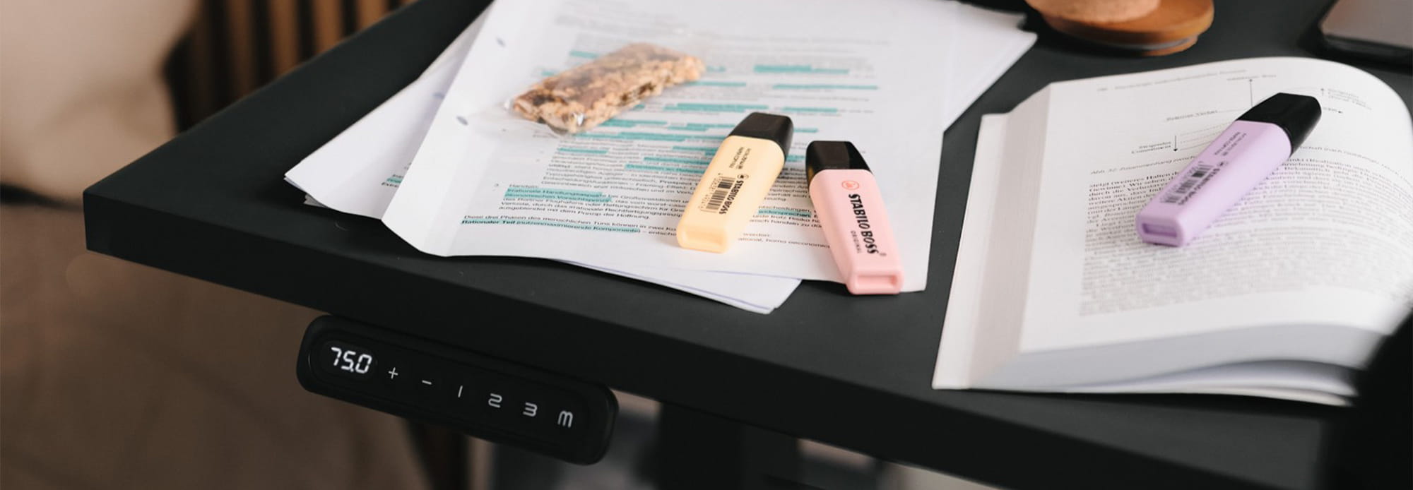 Height-adjustable desk with control panel showing "75.0", with paper, highlighters, and an open book on it.