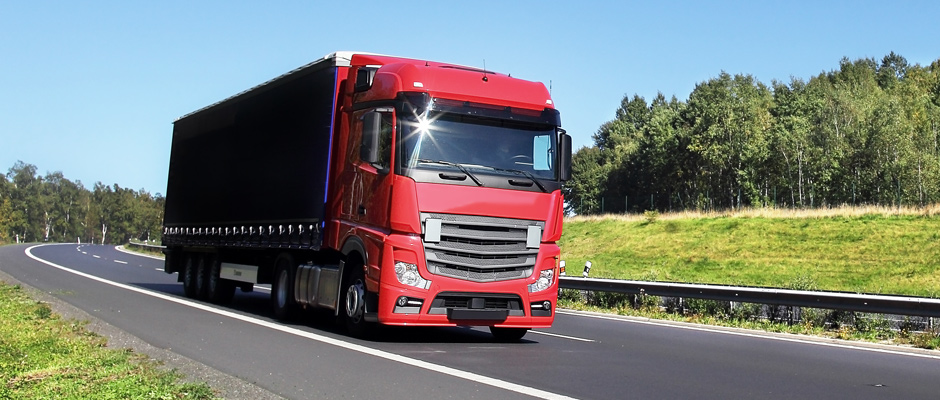 Roter Lastwagen fährt auf einer Straße entlang, umgeben von grünen Feldern und Bäumen.