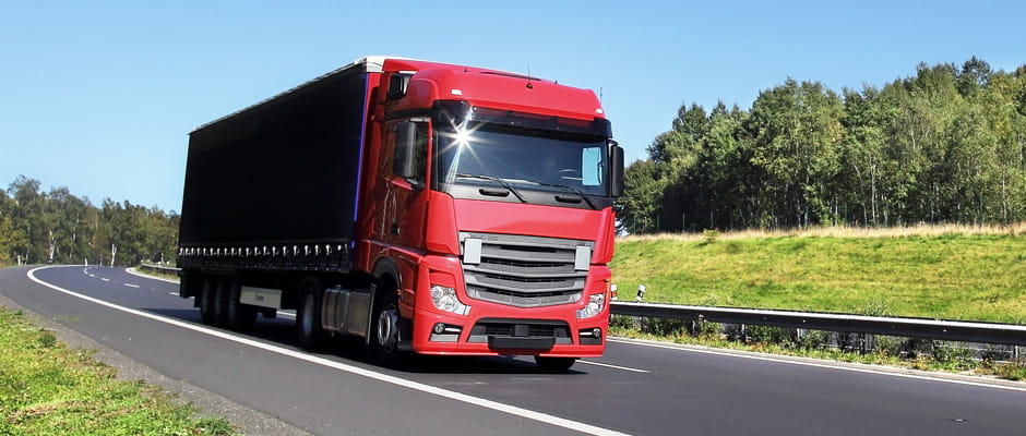 Red truck driving along a road surrounded by green fields and trees.