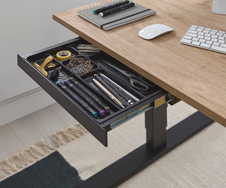 Bureau assis-debout avec un plateau en bois et un cadre noir, doté d’un tiroir sous le bureau ouvert, contenant soigneusement des fournitures de bureau telles que des stylos, des ciseaux et des trombones.