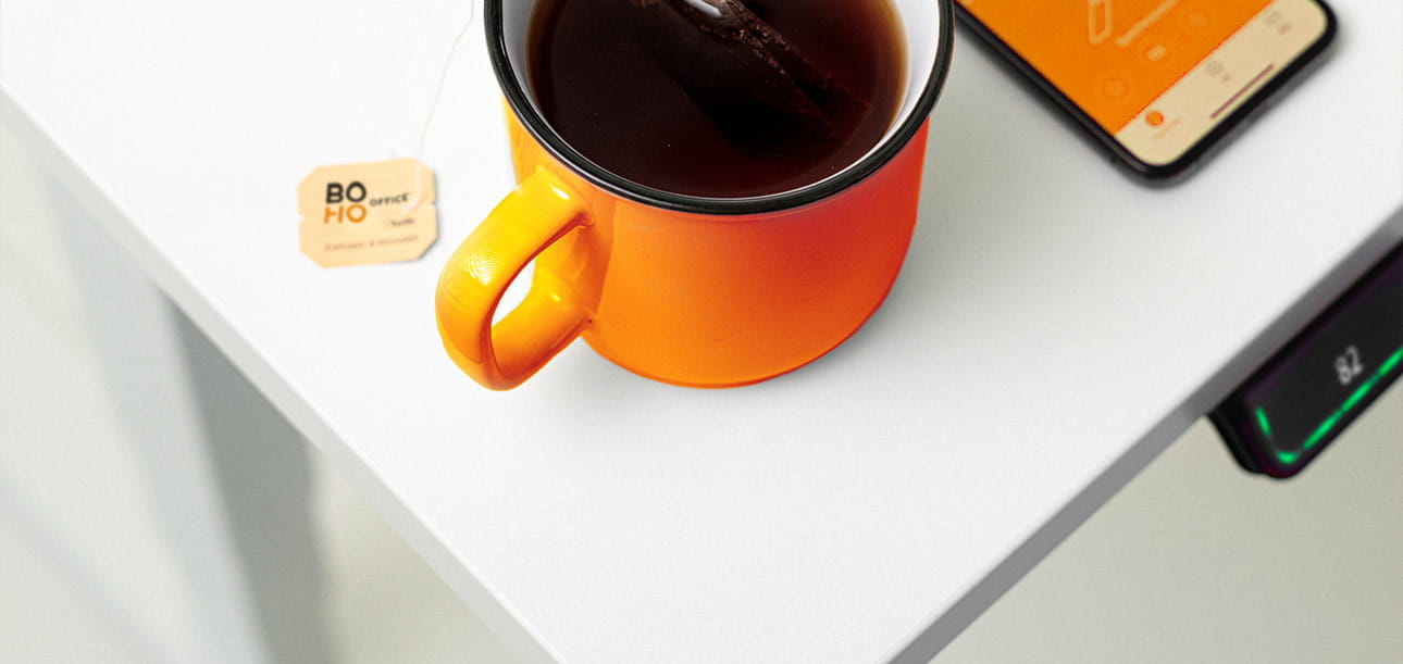 Orangefarbene Tasse mit Kaffee und Teebeutel auf einem weißen Schreibtisch, Smartphone und Bedienelement für die Höhenverstellung sichtbar.