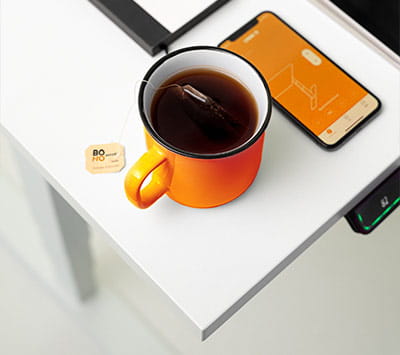 Eine orangefarbene Tasse mit Kaffee und einem Teebeutel auf einem weißen Schreibtisch. Ein Smartphone und ein Bedienelement für die Höhenverstellung sind sichtbar.