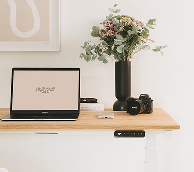 Ein Schreibtisch mit Holzplatte, darauf ein Laptop, eine Kamera, eine Vase mit Blumen, ein Kerzenhalter und ein Magazin. 