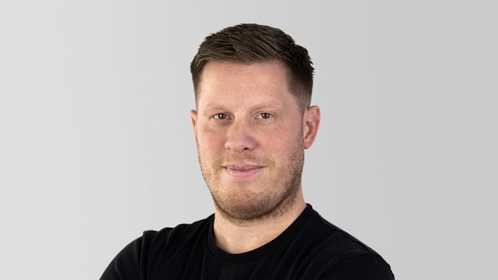Junger Mann mit 3-Tage-Bart lächelt in die Kamera vor neutralen Hintergrund.