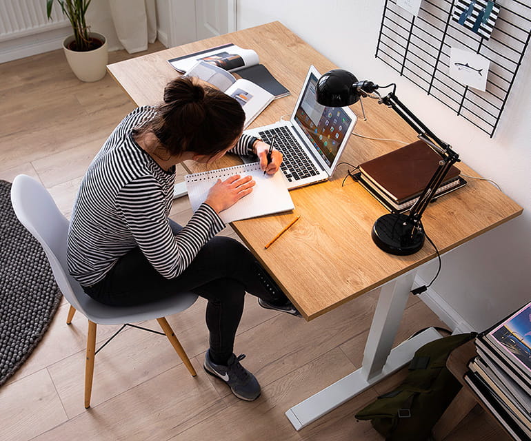 Frau sitzt an einem höhenverstellbaren Schreibtisch mit Holzplatte und weißem Gestell, arbeitet in einem Notizbuch neben einem Laptop, einer Schreibtischlampe und Büchern in einem modernen Arbeitszimmer.