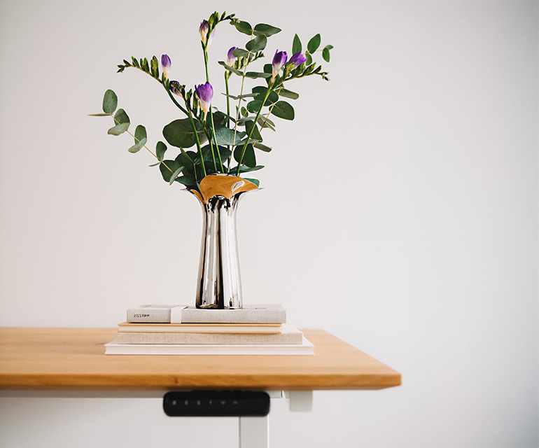 Höhenverstellbarer Schreibtisch mit heller Holzplatte und weißem Gestell, dekoriert mit einem Blumenstrauß in einer Metallvase auf gestapelten Büchern vor einer weißen Wand.