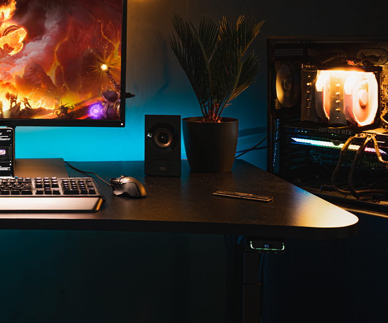 Ein dunkler Raum mit einem Schreibtisch, auf dem ein Monitor und eine Tastatur mit Maus zu sehen sind. Die Tischplatte ist schwarz und zeigt Lichtreflexionen vom einem beleuchteten Computergehäuse im Hintergrund. Sie ist geschwungen mit einem nach innen g