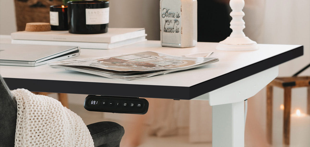 Weißer Schreibtisch mit Laptop, Zeitschriften, Notizbuch, Dekorationsgegenständen wie Kerzen und Vase, Bedienelement für die Höhenverstellung sichtbar.