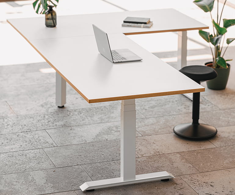 Bureau assis debout moderne en forme de L avec un plateau blanc et un bord en bois, équipé d’un ordinateur portable et de plantes de bureau dans une pièce lumineuse.