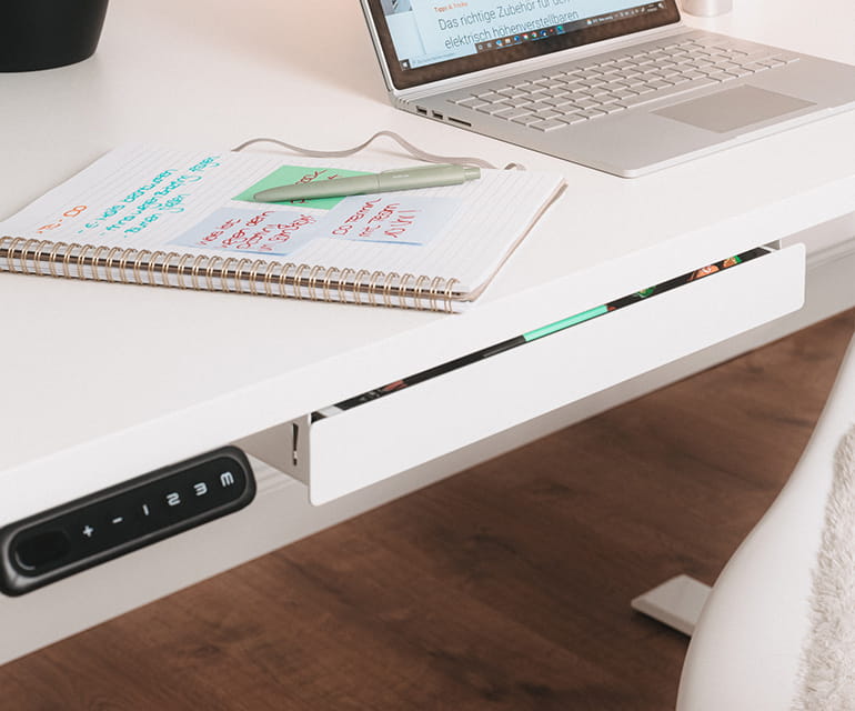 Bureau assis-debout blanc avec un tiroir intégré sous le bureau, partiellement ouvert, à côté d’un panneau de commande numérique, avec un ordinateur portable et un carnet sur le plateau.