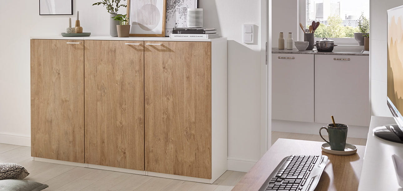 Schrank mit Holzfronten und weißen Seiten, Computer, Tasse und Tastatur auf dem Tisch, Küche im Hintergrund.