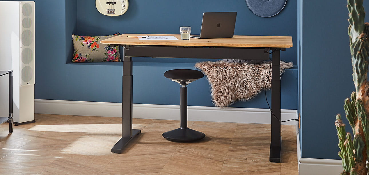 Höhenverstellbarer Schreibtisch mit Holzplatte, Laptop und Glas Wasser, Steh-Sitzhocker daneben, Wanddekoration im Hintergrund.