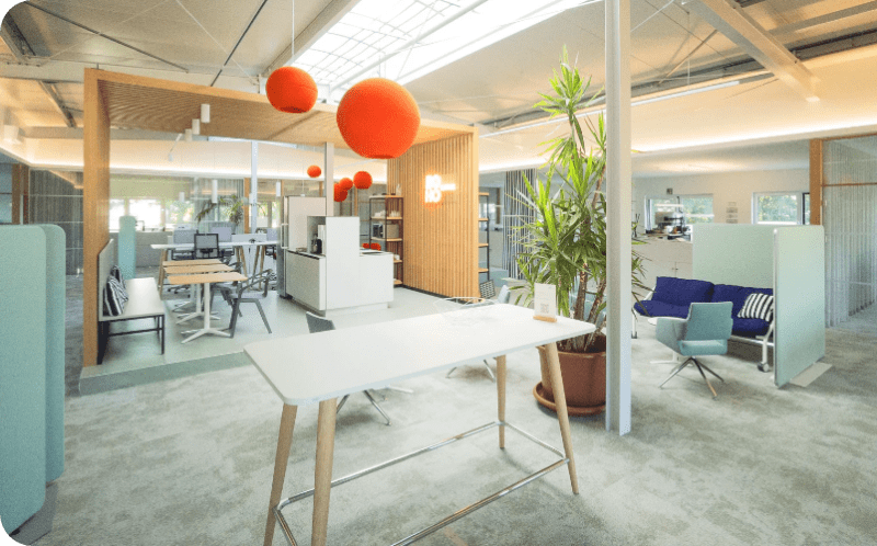 Modern office with a table in the foreground, office workstations and plants in the background. Three orange spheres hang from the ceiling.