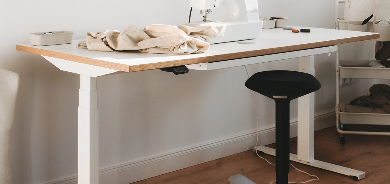 Height-adjustable desk with wooden top, sewing machine, fabrics, and standing seat aid (standing stool), additional furniture in the background.