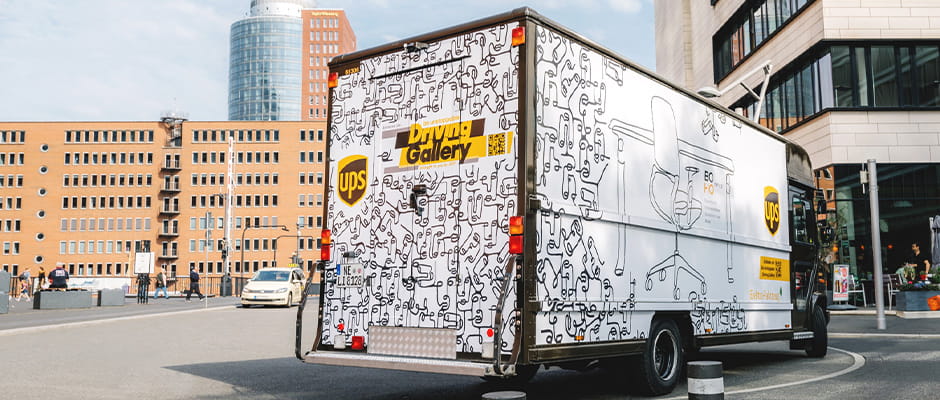 UPS delivery van with artistic graphics driving on a city street, surrounded by modern buildings.