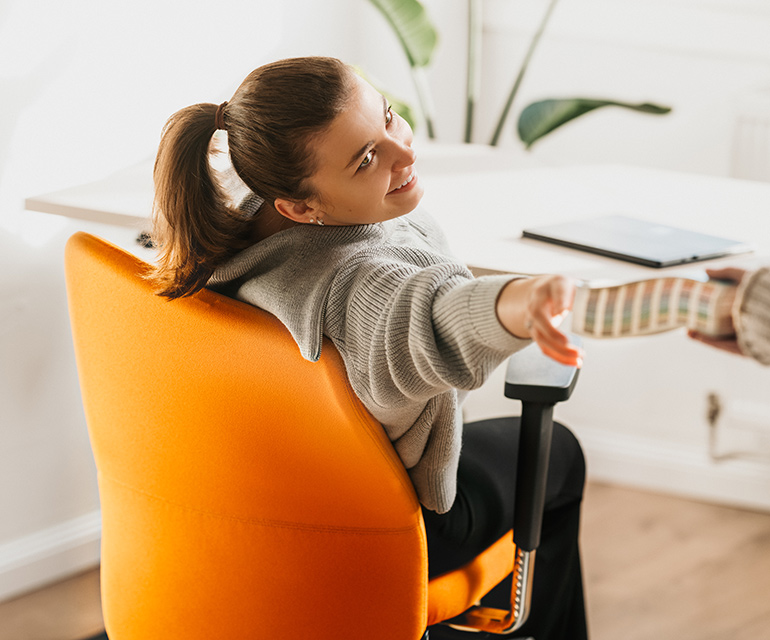 Person sitzt auf einem ergonomischen Bürostuhl mit orangefarbenem Polster, dreht sich lächelnd um und greift nach einer Farbpalette, die ihr überreicht wird.