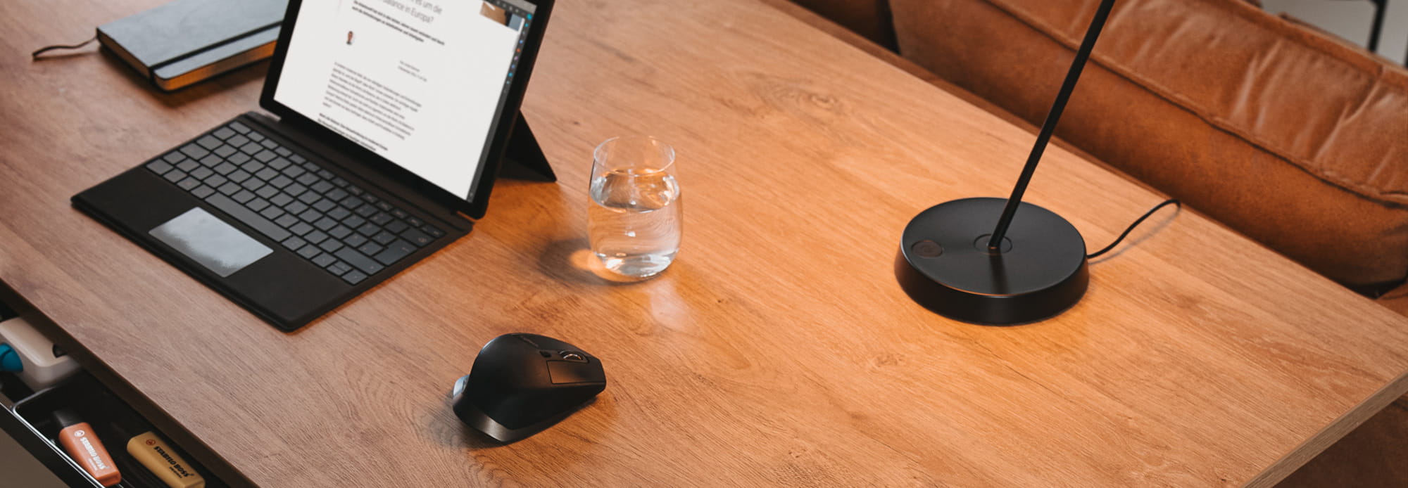 Holz-Schreibtisch mit Laptop, Maus, Glas Wasser und Lampe, minimalistische Ausstattung.