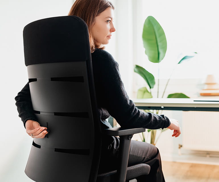 Osoba siedząca na ergonomicznym krześle biurowym, regulująca oparcie za pomocą suwaka, z biurkiem w tle.