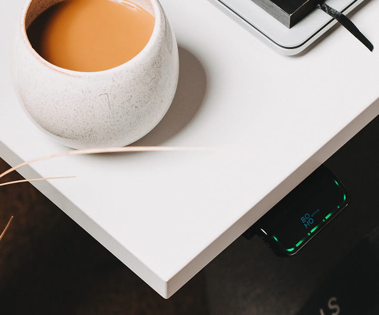 Die Ecke einer weißen Tischplatte mit der Bedieneinheit eine elektrisch höhenverstellbaren Schreibtischs. Auf der Schreibtischplatte sieht man eine Kaffeetasse mit einem Milchkaffee und einen Notizblock.