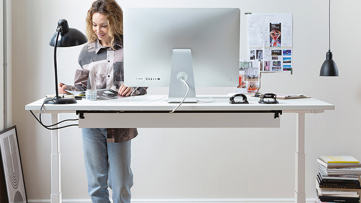 Frau arbeitet an einem weißen höhenverstellbaren Schreibtisch im Stehen, ausgestattet mit einem großen Monitor, einer Schreibtischlampe und Organisationszubehör.