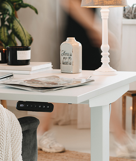 Weißer Schreibtisch mit Lampe, Behälter, Zeitschriften und Pflanze. Bedienfeld am Tisch, Person im Hintergrund in Bewegung.