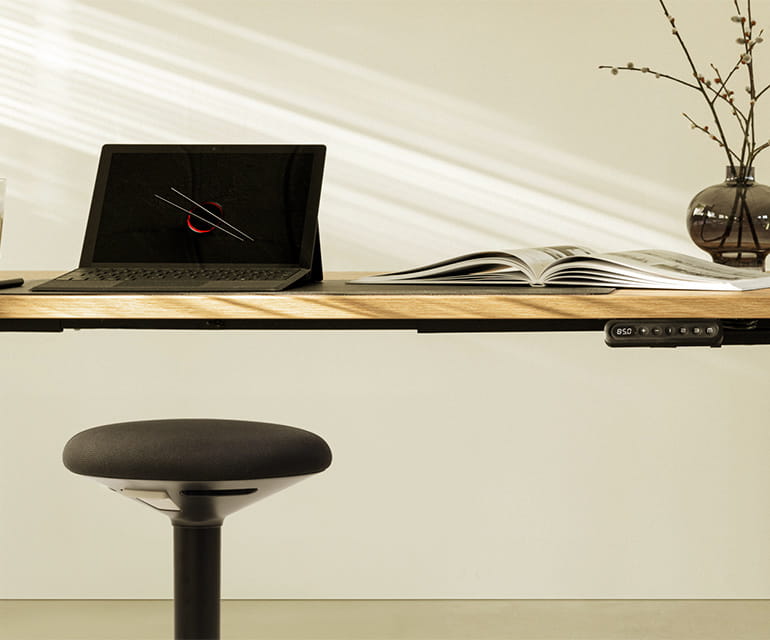 Höhenverstellbarer Schreibtisch mit Holzplatte, ausgestattet mit einem digitalen Bedienfeld, einem Laptop, einem offenen Buch und einer Vase mit Zweigen, dazu ein ergonomischer Hocker.