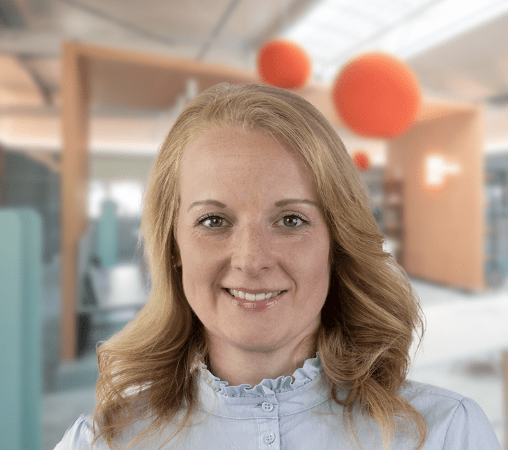 A woman with blonde hair and a friendly smile, in front of a modern office background with orange lamps.