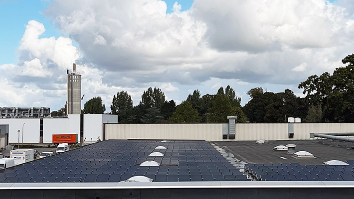 Photovoltaik-Anlage auf Industriedach, tagsüber, Wolken im Himmel, grüne Bäume im Hintergrund.