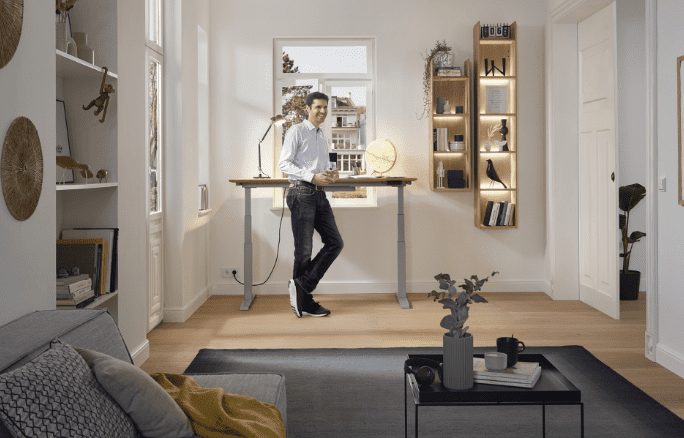 Mann steht an höhenverstellbarem Schreibtisch mit Lampe und Globus in modernem, lichtdurchflutetem Raum.