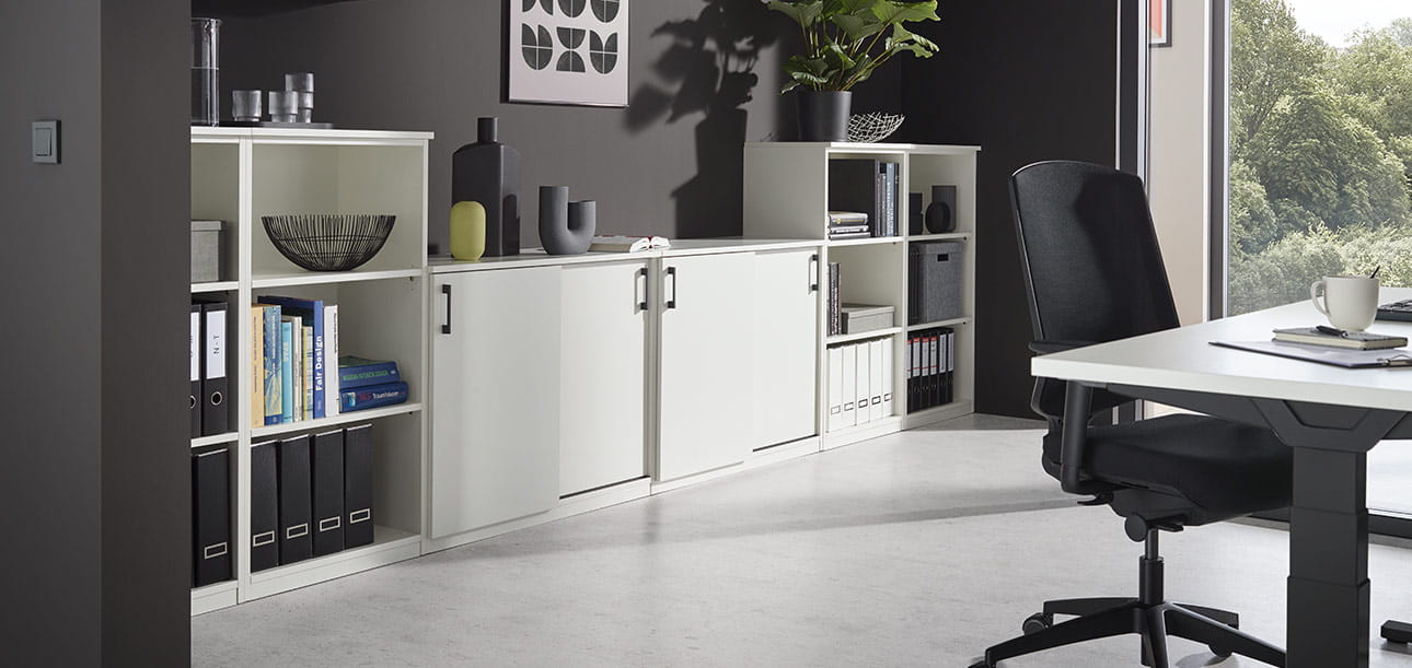 Moderner Bürobereich mit Regalen und Schränken voller Ordner und Dekor, Schreibtisch mit Stuhl im Vordergrund, große Fenster lassen Tageslicht herein.