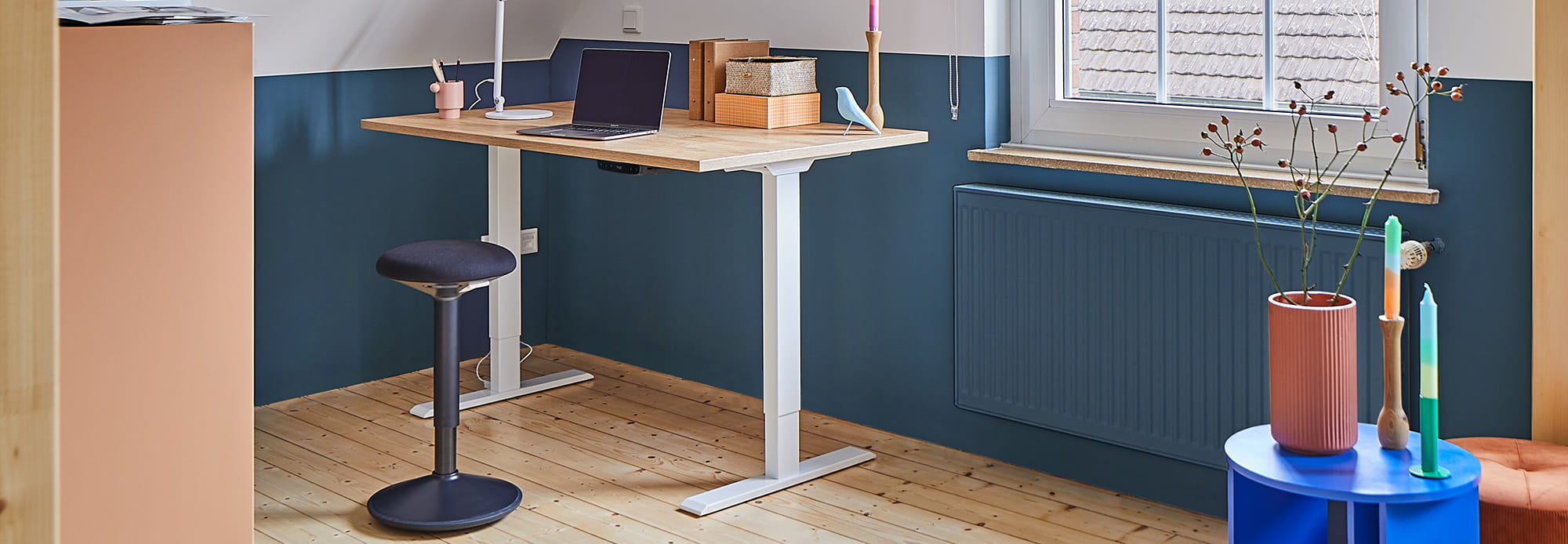 Höhenverstellbarer Tisch mit Laptop und Schreibtischutensilien, Stehhocker, Pflanzen und Dekorationsgegenstände in einem Raum mit Holzfußboden und bunter Wand.