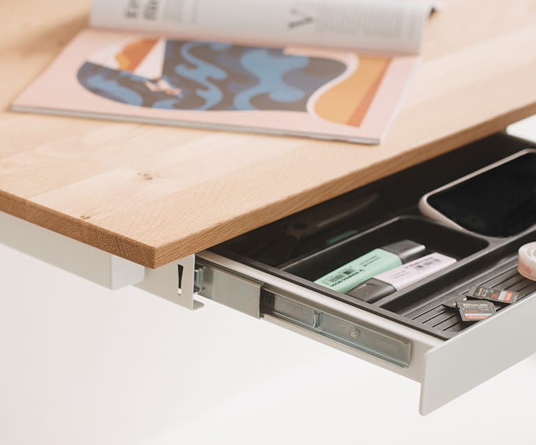 Bureau assis-debout blanc avec un plateau en bois, doté d’un tiroir sous le bureau ouvert, contenant soigneusement des fournitures de bureau telles que des surligneurs, des cartes mémoire et des trombones.