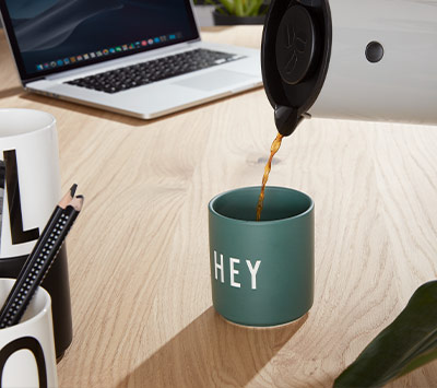 Eine Tasse mit dem Aufdruck "HEY" wird mit Kaffee aus einer Kanne gefüllt. Auf dem Tisch befinden sich ein Laptop, Stifte und eine Pflanze.