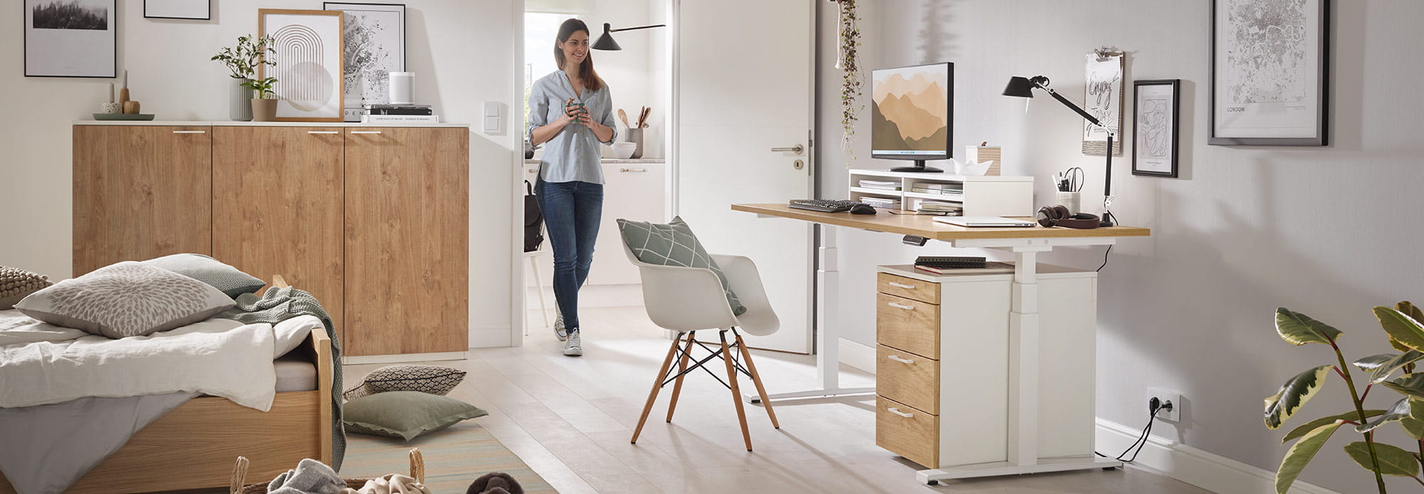 Modern office with height-adjustable desk, chair, and desk supplies. A person enters the room, bed and wardrobe in the background.
