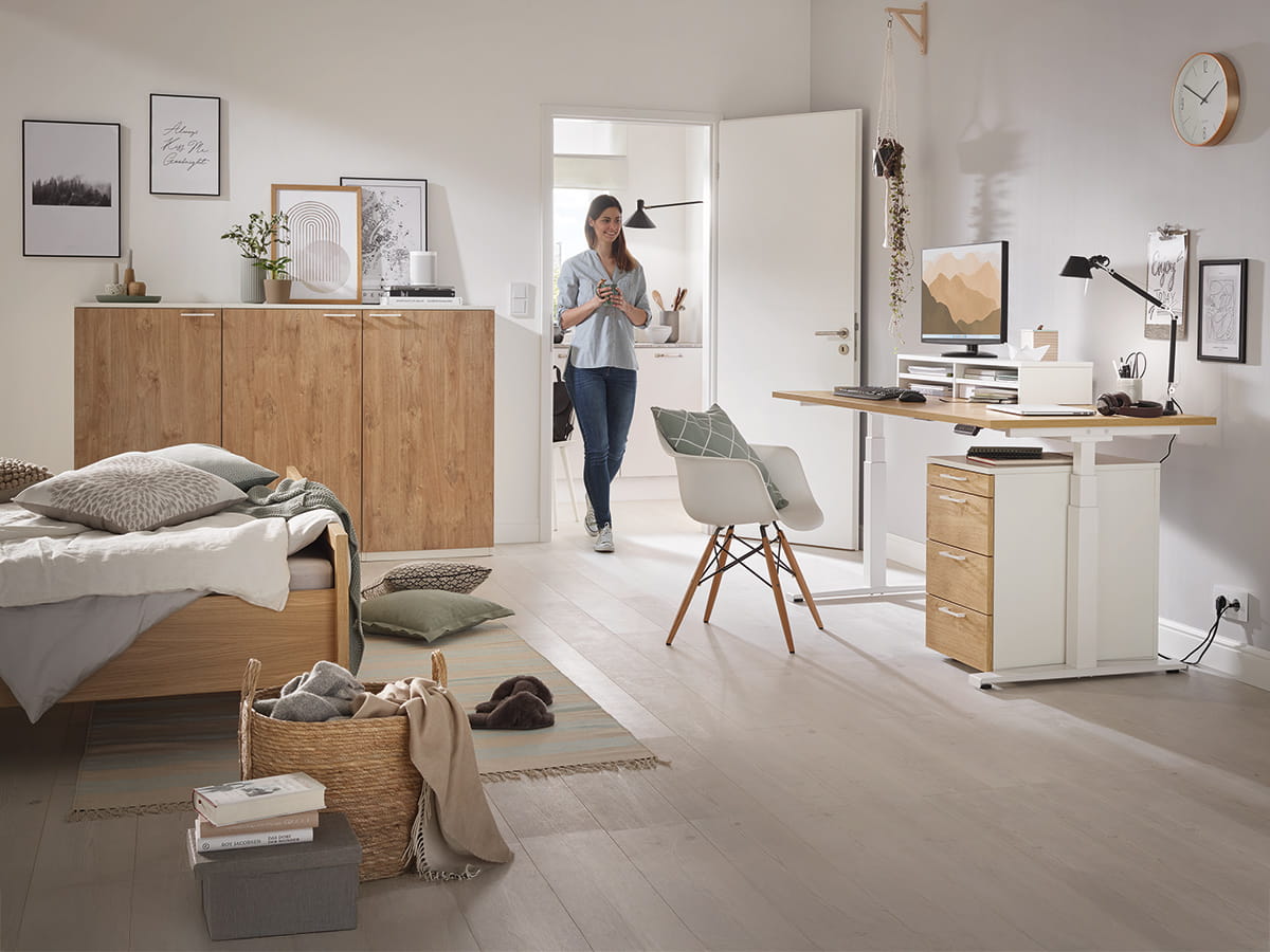 Ein moderner Raum mit Bett, Holzschrank und einer stehenden Person im Türrahmen. Büroeinrichtung am Schreibtisch.