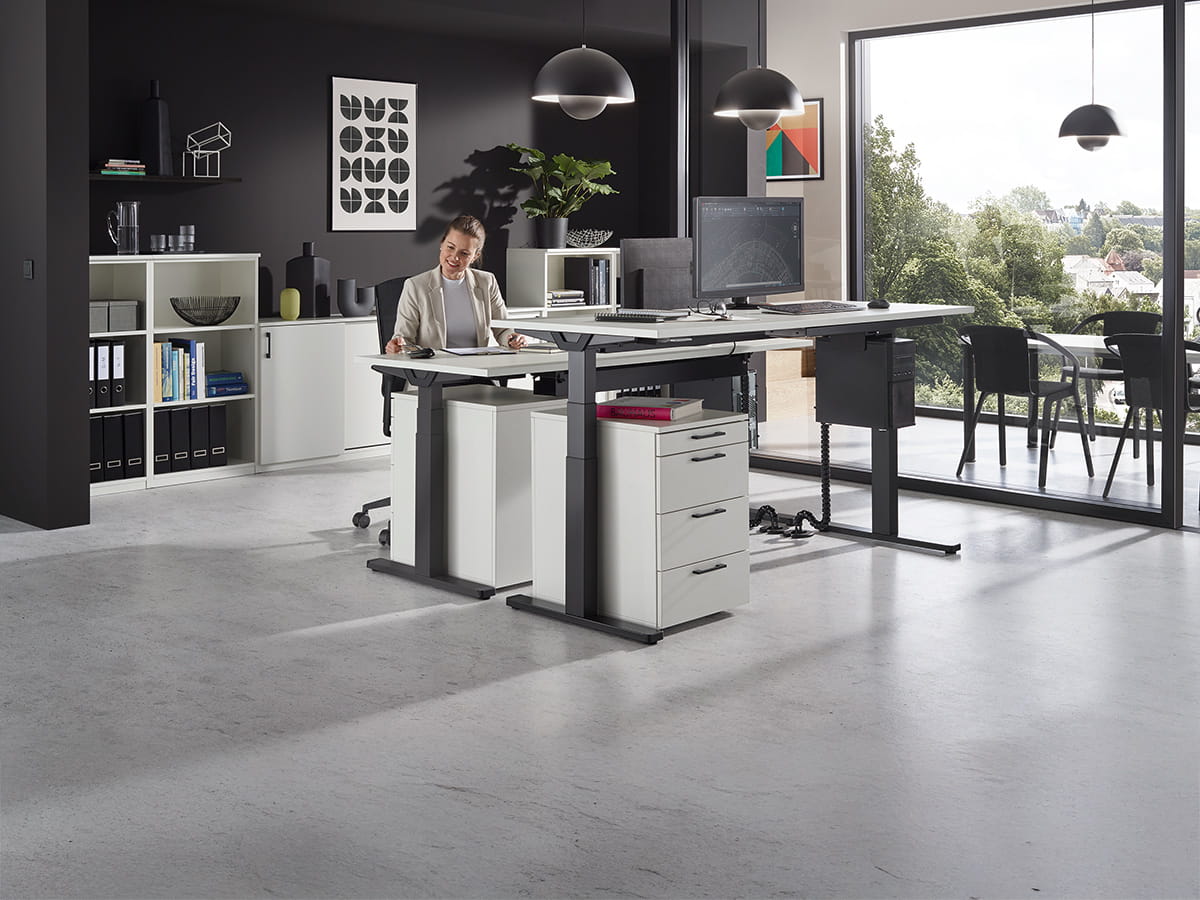 Modernes Büro mit höhenverstellbarem Schreibtisch, Rollcontainer, Regalen und großer Fensterfront mit Ausblick.