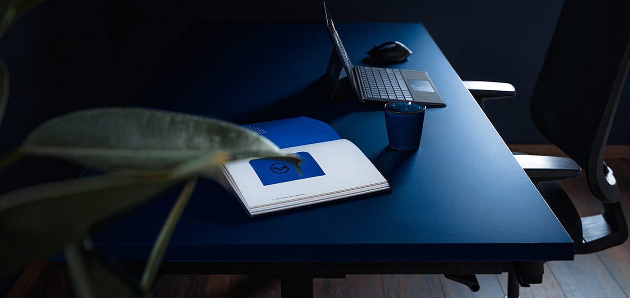 Blauer Schreibtisch mit Laptop, Notizbuch und Glas, Bürostuhl im Hintergrund.