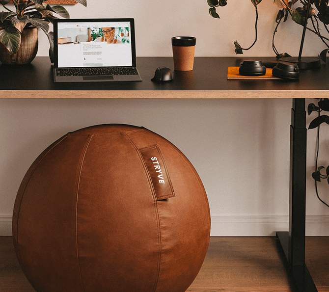 Schreibtisch mit Laptop, Kaffeebecher, Pflanzen und braunem Sitzball in modernem Büro mit Holzfußboden.