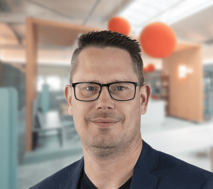 Ein Mann mit Brille und kurzem, dunklem Haar, der in die Kamera lächelt, vor einem modernen Bürohintergrund.